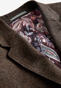Blazer in tweed marrone con un'esterno testurizzato e una fodera interna a fantasia che presenta disegni floreali e paisley in borgogna, verde e bianco.
