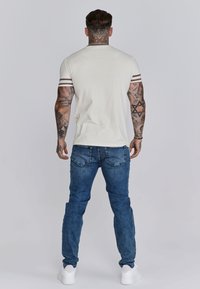 Heren lichtcrème t-shirt met wijnrode strepen op de mouwen, gecombineerd met medium blauwe denim jeans en witte sneakers.