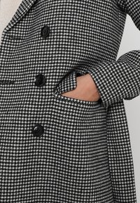 Kabát s černo-bílým houndstooth vzorem, dvouřadý design, s černými knoflíky a boční kapsou, vyrobený z texturované tkaniny.