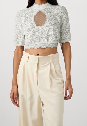 Kvinde iført en hvid, tekstureret cropped top med korte ærmer og en rund netudskæring på brystet samt højtaljede cremefarvede plisserede bukser.