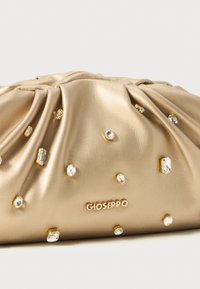 Beige laukku, jossa on rypytystä ja joka on koristeltu erikokoisilla kirkkailla paineteillä sekä metallisella "GIOSEPPO"-logolla etuosassa.