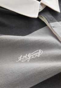 Polo shirt featuring horizontal stripes in gray and black, with a white collar. Includes a white signature printed on the fabric.