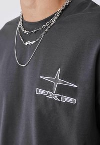 Camiseta de algodón gris con logo bordado en blanco "PXP" y diseño de estrella en el lado izquierdo del pecho, llevada con cadenas de plata en capas alrededor del cuello.