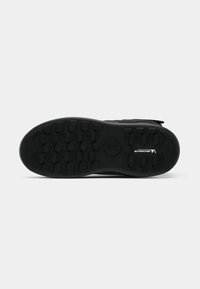 Semelle de chaussure noire en caoutchouc texturé, présentant des motifs de semelle hexagonaux pour une adhérence et un logo subtil au centre. Finition lisse sur les bords.