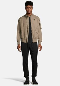 Beige bomberjack met een ritssluiting, geribde manchetten en zakken. G gedragen over een zwart shirt en gecombineerd met zwarte cargo-pants en sneakers.