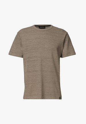 Uomo che indossa una T-shirt beige a maniche corte con scollo rotondo, in piedi davanti a uno sfondo chiaro uniforme, guardando leggermente verso il basso.