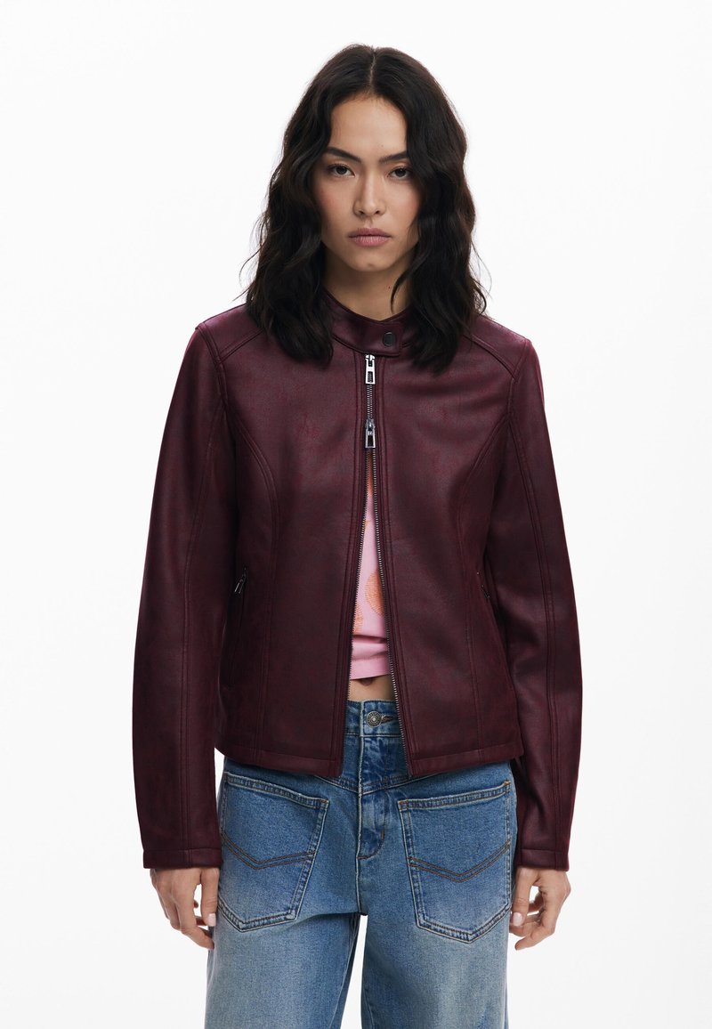 Giacca in pelle bordeaux con zip frontale, colletto con bottoni e dettagli delle cuciture. Indossata sopra una maglietta rosa e abbinata a jeans in denim blu.