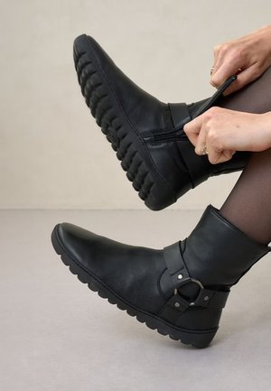 Handen trekken de rits van een zwarte enkellaars met dikke, gestructureerde zool, gedragen met donkere panty's, tegen een effen neutrale achtergrond.