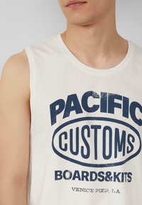 Weißes, ärmelloses Baumwolltanktop mit auffälligem, navyblauem Text: "PACIFIC CUSTOMS BOARDS&KITS" und "VENICE PIER, L.A." im Vintage-Look.