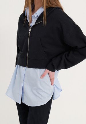 Persona vistiendo una sudadera negra corta con cremallera sobre una camisa azul clara abotonada y pantalones negros, con la mano apoyada en la cadera.