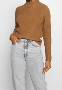 Personne portant un pull en maille marron rentré dans un jean gris clair taille haute, une main dans la poche, sur un fond uni.