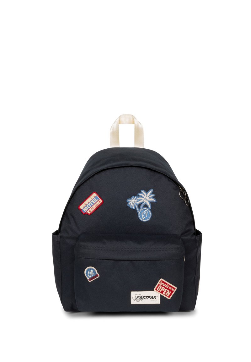 Mochila de un oscuro color navy con forma redondeada, material de tela, múltiples parches bordados y un bolsillo frontal con cremallera. Cuenta con un asa en la parte superior y correas ajustables.