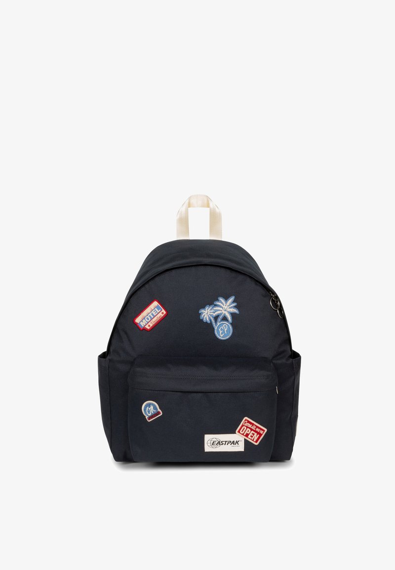 Mochila de un oscuro color navy con forma redondeada, material de tela, múltiples parches bordados y un bolsillo frontal con cremallera. Cuenta con un asa en la parte superior y correas ajustables.