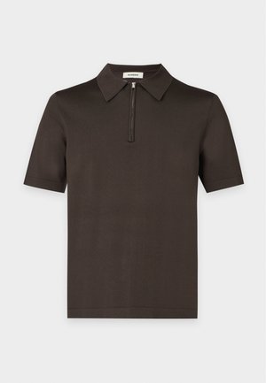 Mann steht und trägt ein dunkelgrünes kurzärmeliges Polohemd, lockere dunkelblaue Hosen und weiße Turnschuhe vor einem einfarbigen Hintergrund.