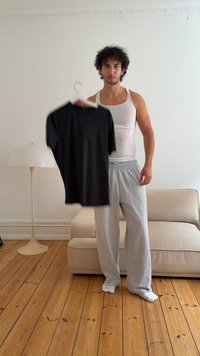 Czarna koszulka z krótkim rękawem zawieszona na wieszaku, wyeksponowana na jasnym tle. Model w szarych spodniach dresowych i białym podkoszulku.