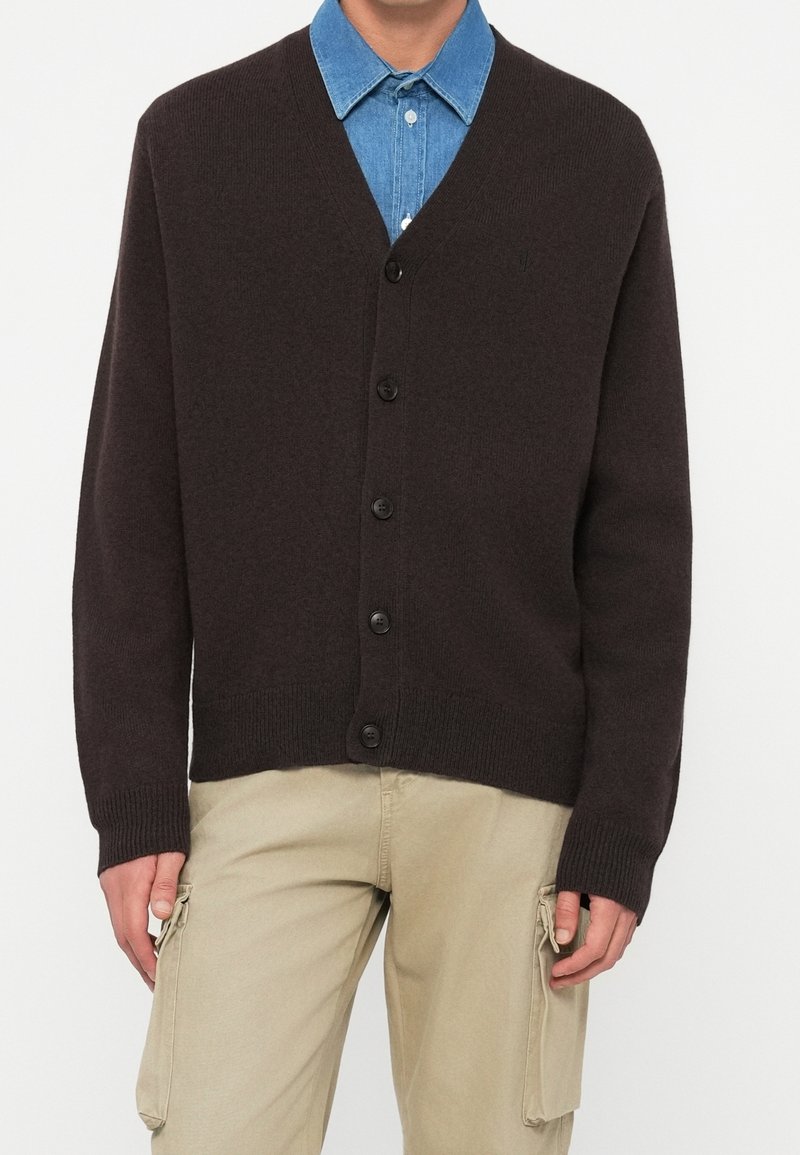 Homme portant un cardigan marron foncé boutonné sur une chemise bleue à col et un pantalon cargo beige.