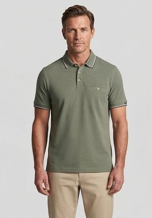 Man staat frontaal, draagt een olijfgroen poloshirt met beige broek tegen een effen lichte achtergrond.