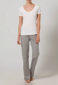 Kvinne som står barfot iført en enkel hvit t-skjorte med korte ermer og løse grå bukser foran en nøytral bakgrunn.