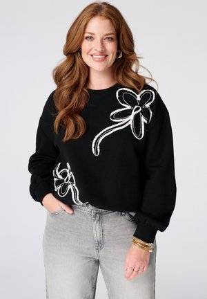 Vrouw met lang rood haar die een zwarte trui met witte bloemmotieven en lichtgrijze spijkerbroek draagt, glimlachend met hand in zak.