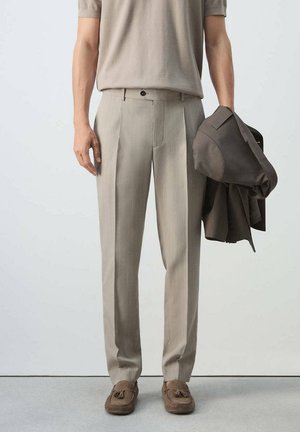 Hombre con pantalones plisados beige, mocasines marrón claro, sosteniendo una chaqueta marrón oscuro en la mano derecha sobre un fondo liso.