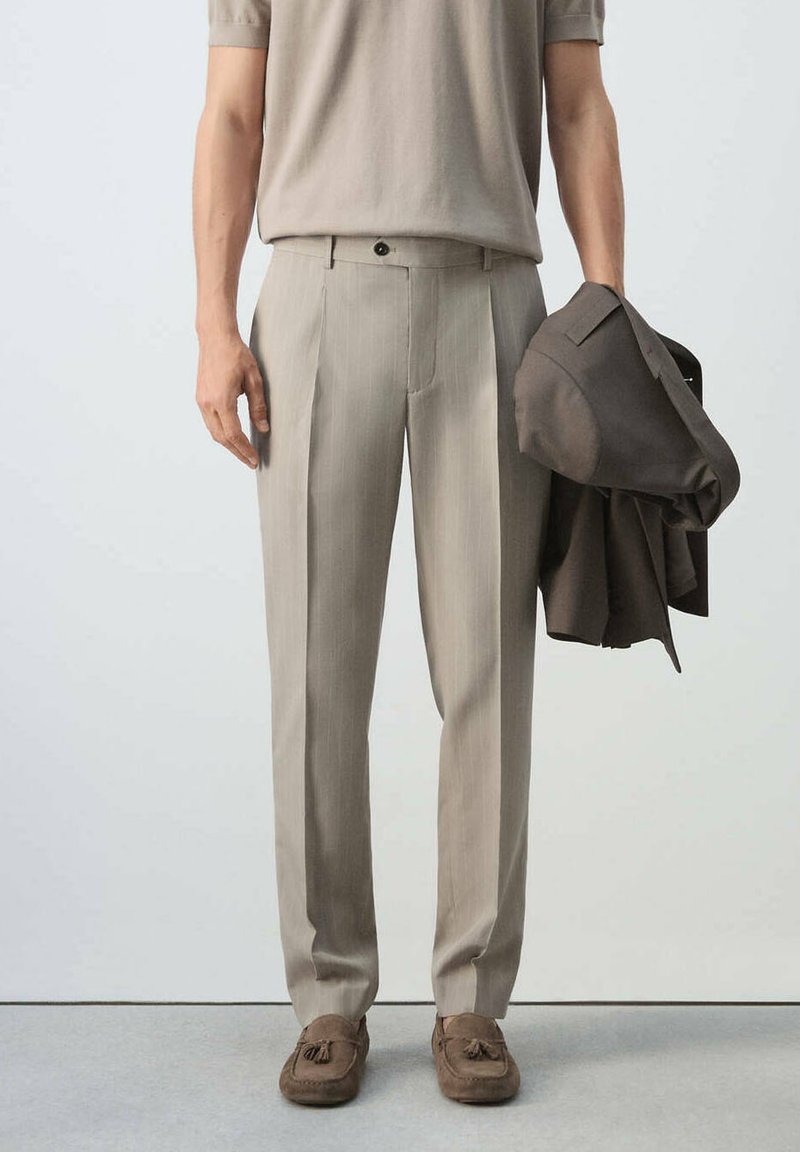 Homme portant un pantalon plissé beige, des mocassins marron clair, tenant une veste marron foncé dans la main droite sur un fond uni.