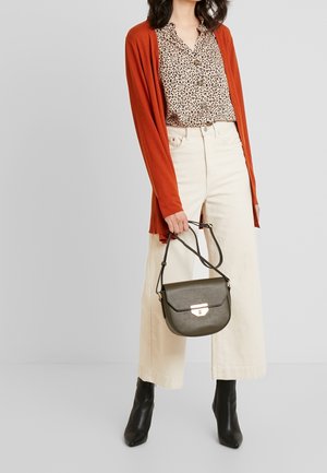 Frau in cremfarbigen weitem Hosenanzug, Bluse mit Leopardenmuster, rostfarbigem Cardigan, schwarzen Stiefeletten, die eine dunkelgrüne Handtasche mit goldener Schnalle hält.