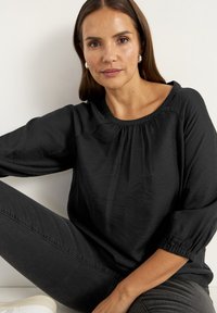 Svart långärmad topp med samlad halslinning och textilstruktur. Bärs tillsammans med mörka jeans; har en avslappnad passform och elastiska ärmslut.