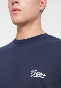 Acercamiento de una persona que viste una camiseta de cuello redondo color azul marino con el texto blanco "Guess Est. 1981 Los Ángeles, CA" en el pecho.