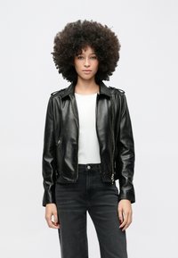 Fiatal nő göndör hajjal, fekete bőrdzsekit, fehér inget és fekete farmert viselve, sima világos háttér előtt állva.