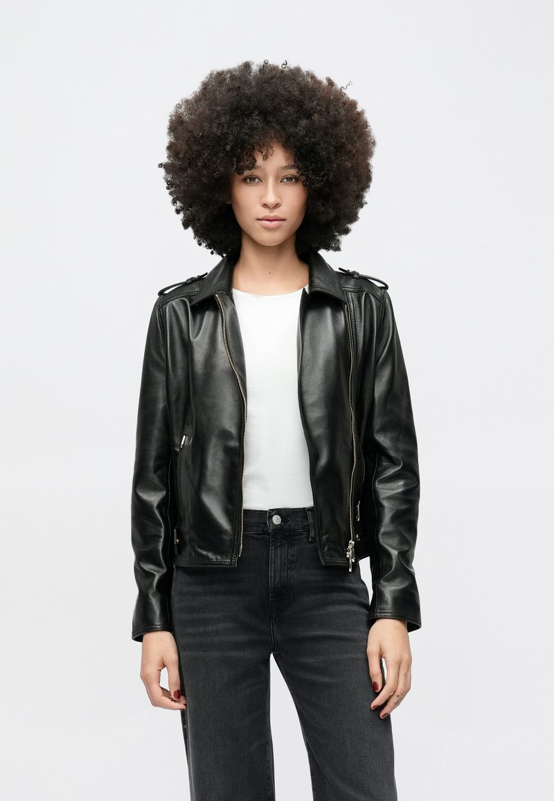 Fiatal nő göndör hajjal, fekete bőrdzsekit, fehér inget és fekete farmert viselve, sima világos háttér előtt állva.