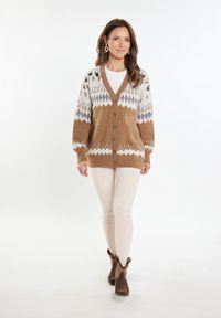 Cardigan a fantasia marrone e bianco con scollo a V, 5 bottoni e maniche lunghe, indossato sopra una maglietta chiara e abbinato a pantaloni beige e stivali marroni.