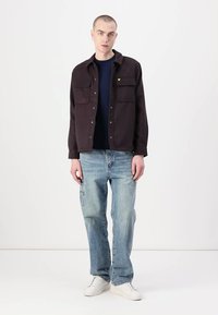 Veste chemise sombre à boutons avec deux poches poitrine, associée à un jean bleu clair et des baskets blanches ; mannequin avec les cheveux courts et des boucles d'oreilles.
