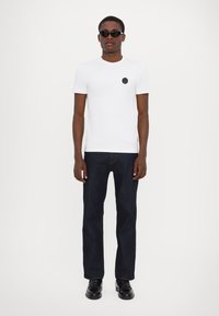Hvid bomulds t-shirt med et sort cirkulært logo på brystet, parret med mørkeblå lige ben jeans og sorte sko. Sorte solbriller bæres.