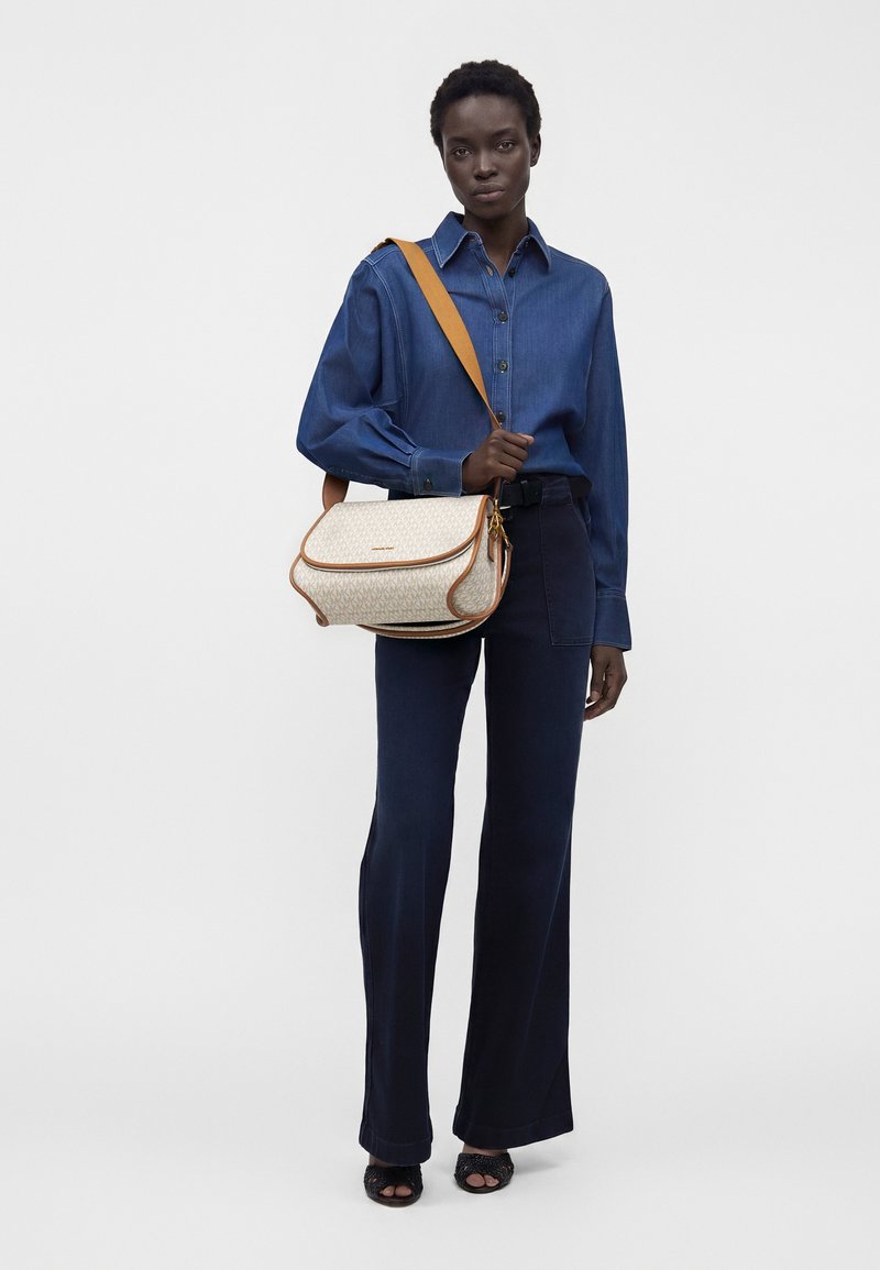 Modèle portant une chemise bleu foncé boutonnée et un pantalon bleu marine avec des sandales à talons noires, portant un sac à épaule beige à motif avec une bandoulière marron.