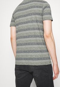 Koszula krótki rękaw w kolorze szarym z geometrycznymi wzorami w zielone i granatowe paski. Materiał ma teksturowaną fakturę i luźny krój.