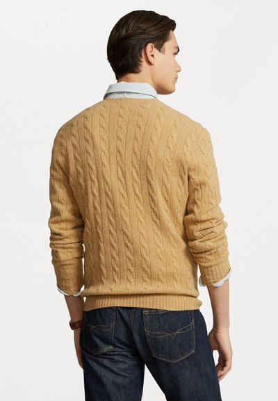 Pulôver de malha trançada em tons de bege, com decote redondo e punhos canelados. Usado sobre uma camisa listrada azul claro, combinado com jeans escuros.