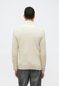 Pull en tricot beige à manches longues, avec un col côtelé, un ourlet droit et une texture douce, porté sur un jean gris, vu de dos.