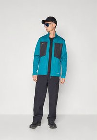 Casaco azul-petróleo com bolsos frontais pretos, t-shirt preta, calças cargo escuras e sapatos pretos; o modelo usa óculos de sol e boné.