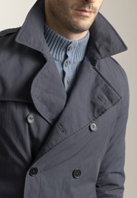 Abrigo trench gris oscuro con un cuello ancho, diseño cruzado y grandes botones negros, sobre un suéter acanalado azul claro.