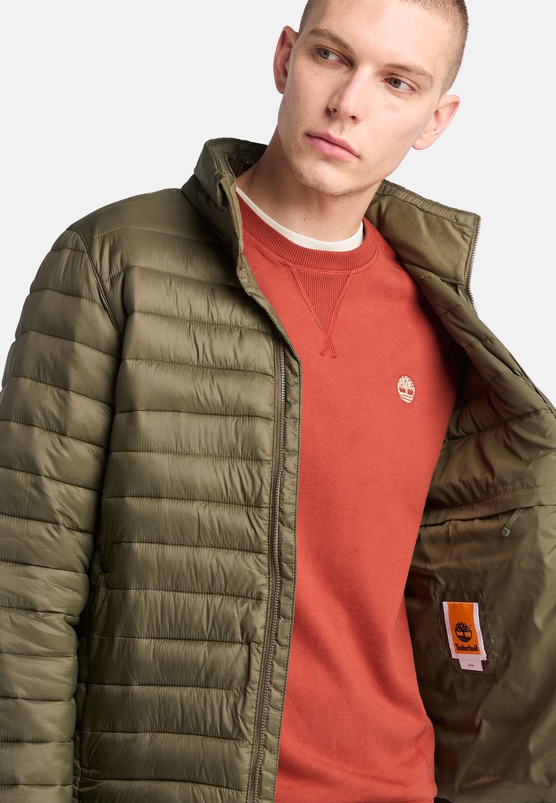 Oliwkowa kurtka puchowa z zamkiem z przodu i prążkowanym kołnierzem, założona na czerwoną bluzę z małym logo.