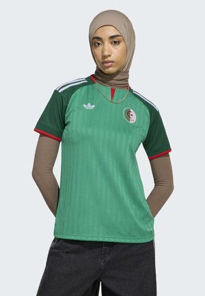 Vrouw die een beige hijab draagt en een gelaagde bruine top met lange mouwen onder een groen Algerijns voetbalshirt met Adidas-strepen en -logo.