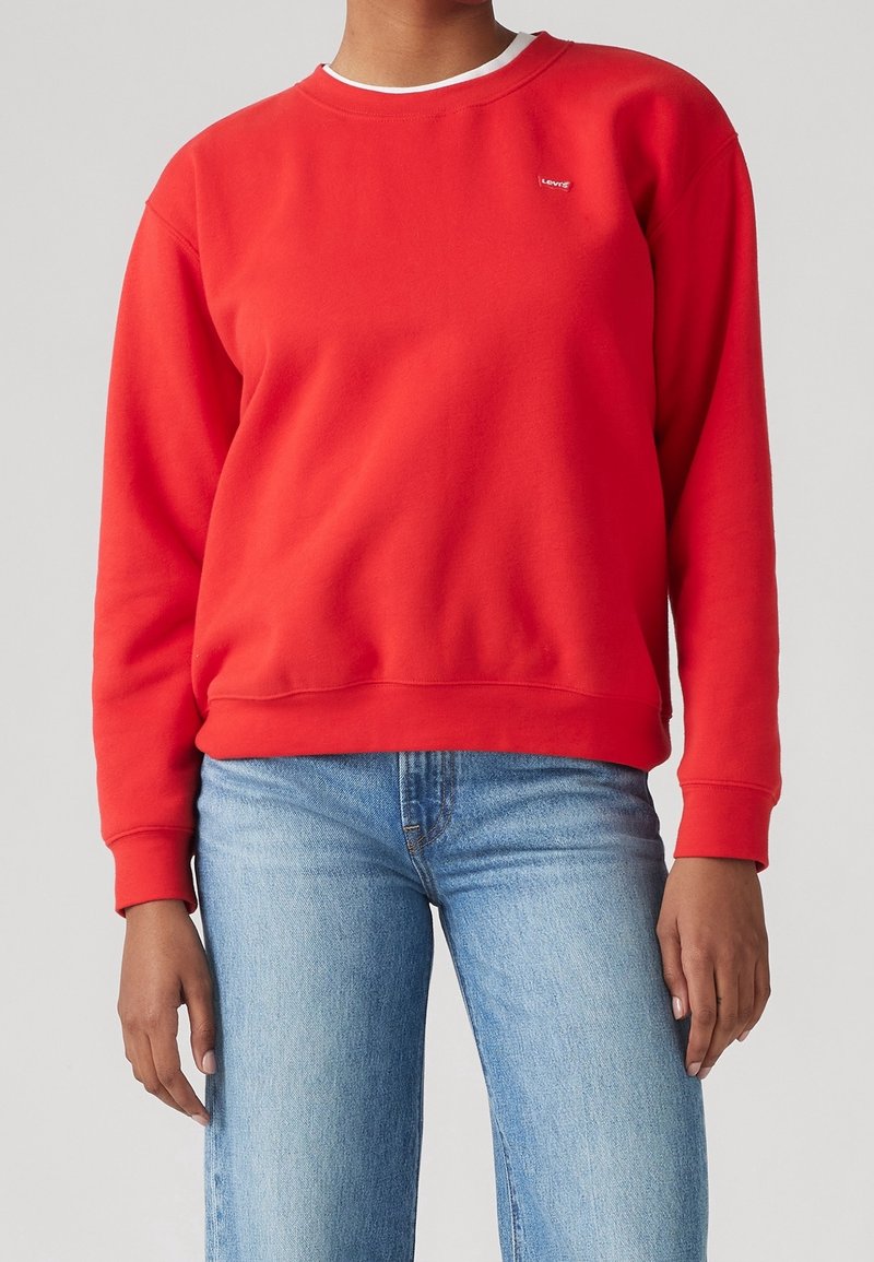Personne portant un sweat-shirt rouge à manches longues avec un petit logo sur la poitrine et un jean bleu clair sur un fond uni.