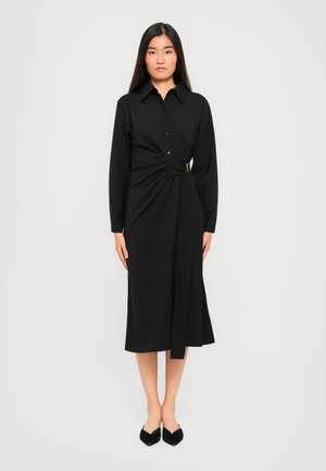 Woman stands wearing a long-sleeve black midi dress with a collar and side buckle detail, paired with black pointed-toe heels on a white background.