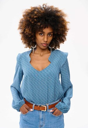 Blauwe blouse met patroon, V-hals, franjeaccenten op de schouders, lange mouwen en een gestructureerde afwerking, gecombineerd met een bruine leren riem.