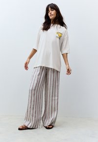 T-shirt oversize crème à manches courtes avec un motif jaune, associée à un pantalon à jambes larges rayé beige et bordeaux. Sandales marron à brides.