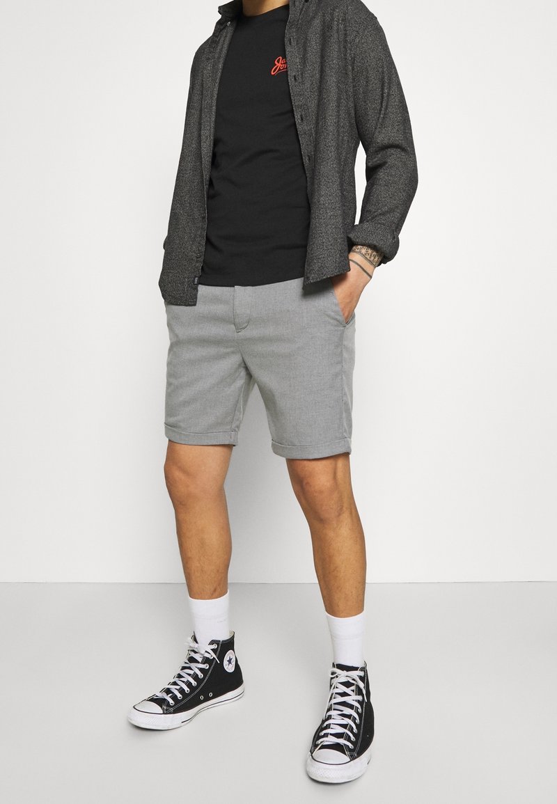 Pantaloni corti grigi con orli arrotolati, abbinati a una maglietta nera e a una camicia botton-down grigio scuro. Sneakers alte nere completano il look.
