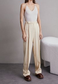 Beige vida byxor med veck, hög midja och skräddarsydd passform. Matchas med en ljusfärgad åtsittande linne och kraftiga bruna skor.
