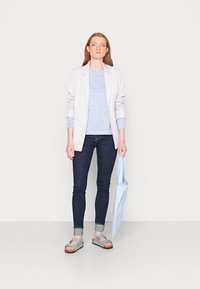 Blazer cinza claro sobre um suéter de malha azul, jeans skinny escuros e sandálias cinzentas. A segurar um guarda-chuva azul com riscas. Design simples.