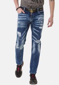 Ausgebleichte blaue Jeans aus Denim mit zerstörten Stellen und Wischdetails. Slim-Fit-Design, kombiniert mit schwarzen Schuhen und einem dunklen Gürtel.