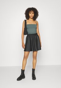 Top ajustado verde escuro com alças finas, combinado com uma saia rodada de imitação de couro preta e botas pretas robustas até o tornozelo. Bolsa preta incluída.